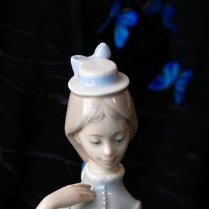 K&ouml;nnte beinhalten: Eine Porzellanfigur einer Person mit hellbraunem Haar, die einen kleinen, hellblauen Hut mit einer Schleife tr&auml;gt. Die Figur hat eine glatte, gl&auml;nzende Oberfl&auml;che und ist ab der Brust aufw&auml;rts dargestellt, mit weichen Gesichtsz&uuml;gen und einem hellblauen Kragen.