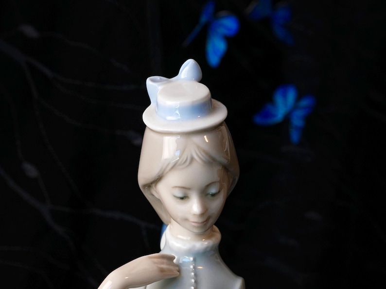 K&ouml;nnte beinhalten: Eine Porzellanfigur einer Person mit hellbraunem Haar, die einen kleinen, hellblauen Hut mit einer Schleife tr&auml;gt. Die Figur hat eine glatte, gl&auml;nzende Oberfl&auml;che und ist ab der Brust aufw&auml;rts dargestellt, mit weichen Gesichtsz&uuml;gen und einem hellblauen Kragen.