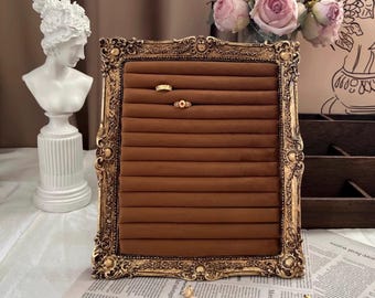 May include: A gold-toned ornate picture frame with a brown velvet interior. The frame has a decorative design and is used to display rings. Two rings are displayed on the velvet.
