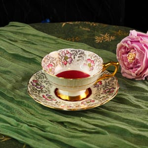 Può includere: Un set vintage di tazza e piattino con un design floreale rosa e oro. La tazza è riempita con un liquido rosso scuro. Il set è posto su un panno verde, con una rosa rosa sullo sfondo.