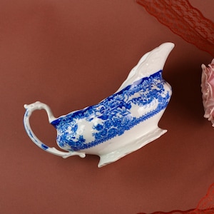 Puede incluir: Salsera de porcelana blanca con un estampado floral azul. El recipiente tiene un asa curva y un pico. El diseño azul y blanco está en el borde exterior e interior. La salsera está sobre una superficie marrón.