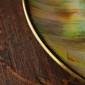 May include: Close-up of a decorative plate with a gold rim. The plate features a colorful abstract design with green, yellow, and brown brushstrokes. The plate is set on a dark wooden surface with inlaid leaf patterns.