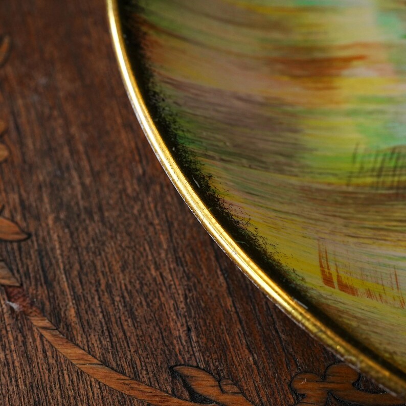 May include: Close-up of a decorative plate with a gold rim. The plate features a colorful abstract design with green, yellow, and brown brushstrokes. The plate is set on a dark wooden surface with inlaid leaf patterns.