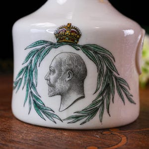 K&ouml;nnte beinhalten: Eine wei&szlig;e Keramikvase mit einem Portr&auml;t eines Mannes im Profil, umgeben von einem gr&uuml;nen Lorbeerkranz. &Uuml;ber dem Portr&auml;t befindet sich eine goldene und juwelenbesetzte Krone. Die Vase hat eine abgerundete Form und einen Henkel.