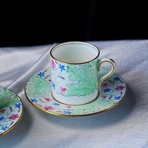 May include: A white porcelain teacup and saucer set featuring a hand-painted botanical pattern of large light green leaves, small pink flowers, and tiny blue blossoms. The rim of the cup and the edge of the saucer are finished with a thin gold trim.
