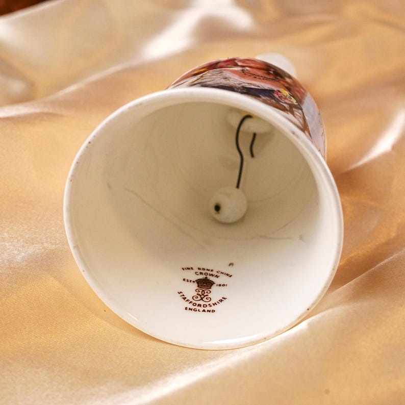 K&ouml;nnte beinhalten: Eine wei&szlig;e Keramikglocke mit einem detaillierten, bemalten Design am oberen Rand. Die Glocke hat einen schwarzen Kl&ouml;ppel und ein dekoratives Emblem am Fu&szlig;. Die Glocke ist aus feinem Knochenporzellan und stammt aus Staffordshire, England.