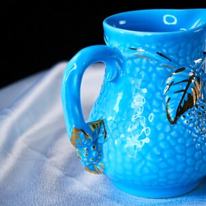 Op de afbeelding: Een fel turquoise keramische kan met een textuur van kleine bobbels. Het handvat en de behuizing zijn versierd met reli&euml;fmotieven van bladeren en bessen die goudkleurig zijn geschilderd. De kan heeft een brede schenktuit en een glanzende afwerking.