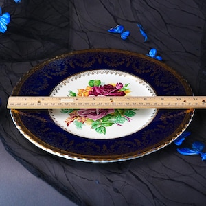 May include: A round porcelain dinner plate featuring a central floral bouquet of red, yellow, and pink roses with green leaves. The plate has a wide, deep blue border with intricate gold filigree detailing. A wooden ruler shows a 10.5-inch diameter.