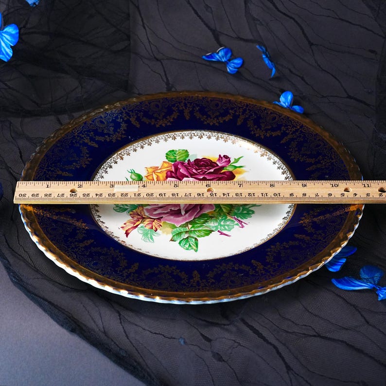 May include: A round porcelain dinner plate featuring a central floral bouquet of red, yellow, and pink roses with green leaves. The plate has a wide, deep blue border with intricate gold filigree detailing. A wooden ruler shows a 10.5-inch diameter.
