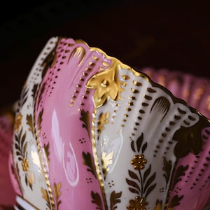 May include: Close-up of a pink and white porcelain teacup with gold leaf accents. The teacup features a scalloped edge and floral designs. The interior is a solid pink, while the exterior has a mix of pink and white sections.