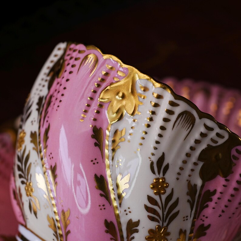 May include: Close-up of a pink and white porcelain teacup with gold leaf accents. The teacup features a scalloped edge and floral designs. The interior is a solid pink, while the exterior has a mix of pink and white sections.