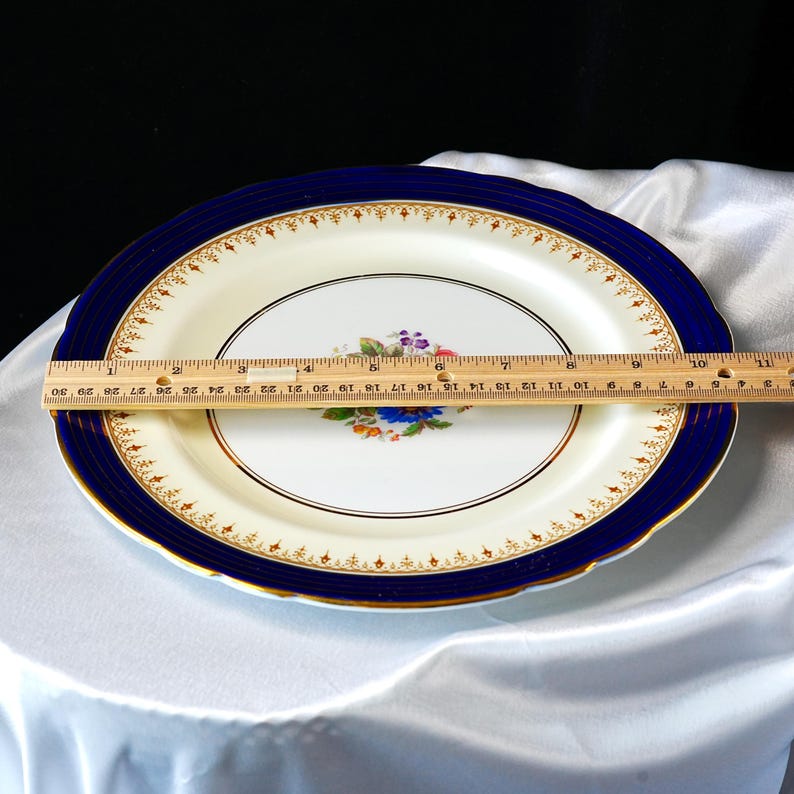 Pode incluir: Uma travessa oval com um centro creme e uma borda azul escura. A borda &eacute; acentuada com detalhes dourados. O centro apresenta um desenho floral. A travessa mede aproximadamente 30 cm de comprimento.