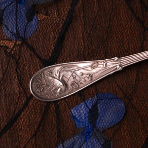 Puede incluir: Un mango de cuchara de metal plateado con un dise&ntilde;o en relieve de un ave en vuelo rodeada de motivos florales. El mango tiene una forma c&oacute;nica con detalles acanalados verticales en el cuello y un extremo redondeado con grabados.