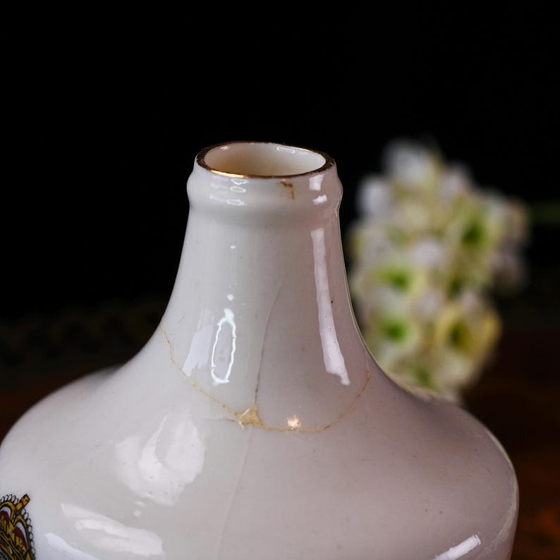 K&ouml;nnte beinhalten: Nahaufnahme einer wei&szlig;en Keramikflasche mit Goldrand. Die Flasche hat einen Riss und ein kleines dekoratives Emblem an der Seite. Der Hintergrund ist dunkel mit einem verschwommenen Blumenarrangement.