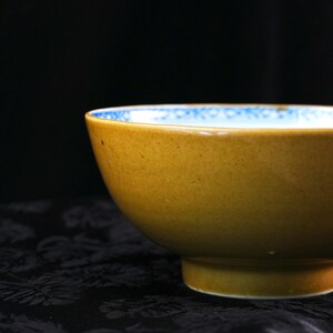 May include: A close-up of a ceramic bowl with a mustard-yellow exterior and a white interior with a blue patterned border. The bowl has a rounded shape and a small, circular base. The background is dark.