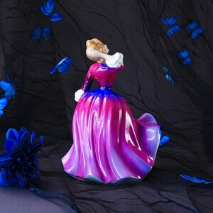 May include: A porcelain figurine of a person wearing a long, flowing gown that transitions from a deep blue at the waist to a vibrant magenta and purple hue at the hem, featuring white accents at the collar and cuffs, viewed from the back.