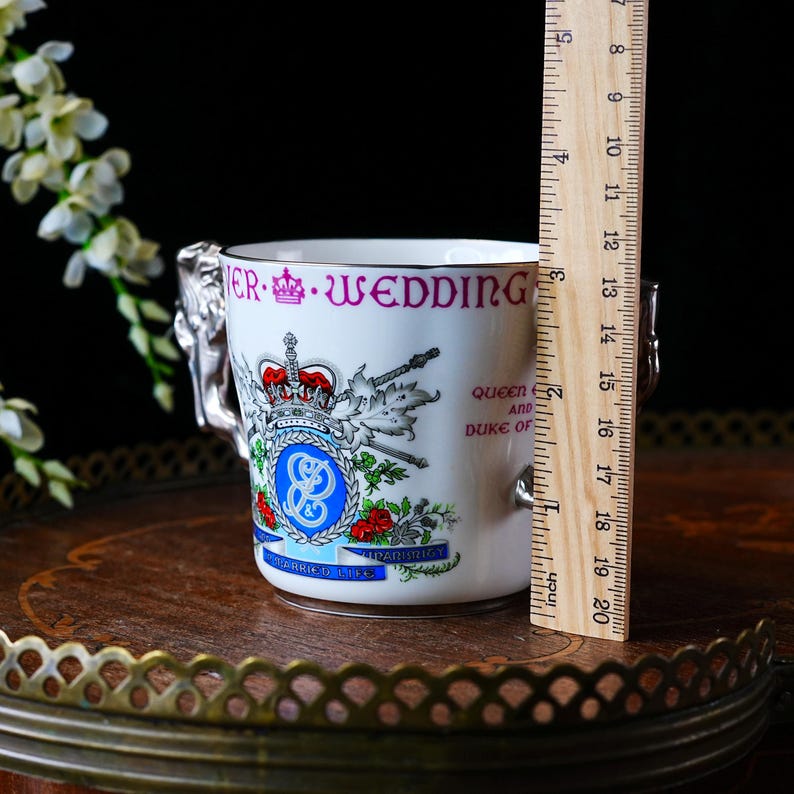 May include: A white commemorative mug with silver handles and a decorative emblem. The mug features the text "ER WEDDING" in purple, along with a royal crest and the words "QUEEN AND DUKE OF". The mug is approximately 7.6 cm tall.