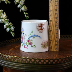 Op de afbeelding: Een witte keramische mok met gouden rand, met illustraties en tekst over een kroning. De mok is ongeveer 10 cm hoog. Decoratieve elementen zijn bloemmotieven en landsemblemen.