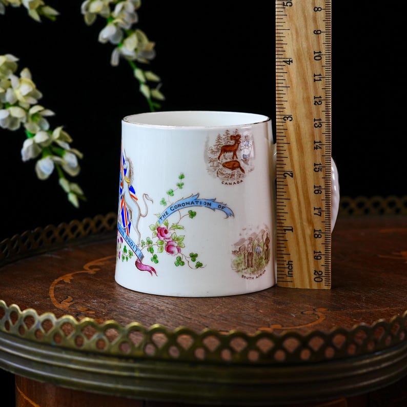 Op de afbeelding: Een witte keramische mok met gouden rand, met illustraties en tekst over een kroning. De mok is ongeveer 10 cm hoog. Decoratieve elementen zijn bloemmotieven en landsemblemen.