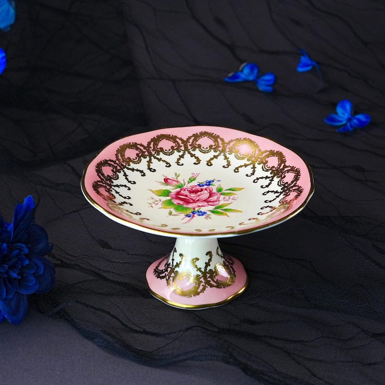 Puede incluir: Un plato de pedestal de porcelana con un centro de color crema con un motivo floral de rosa rosa, rodeado por un borde ancho de color rosa decorado con intrincados patrones de filigrana dorada. La base tambi&eacute;n es rosa con detalles dorados.