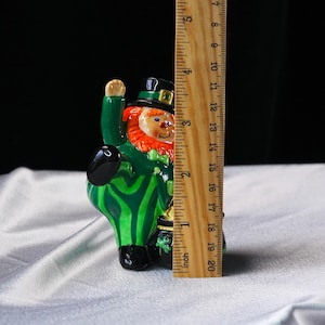 May include: A ceramic leprechaun figurine, approximately 4 inches tall, with a green suit, orange beard, and black hat. The figurine is holding a black pot of gold. The design includes green and black stripes.