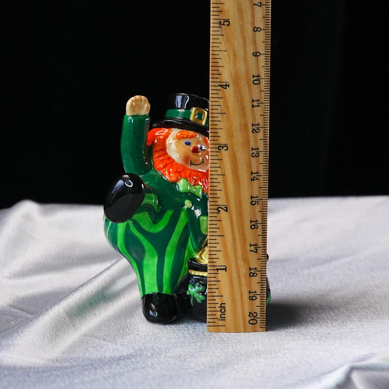 May include: A ceramic leprechaun figurine, approximately 4 inches tall, with a green suit, orange beard, and black hat. The figurine is holding a black pot of gold. The design includes green and black stripes.