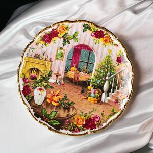 Pu&ograve; includere: Piatto decorativo con una scena natalizia. Il piatto presenta un'illustrazione dettagliata di una stanza con camino, albero di Natale e persone riunite attorno a un tavolo. Il piatto &egrave; rifinito con oro e motivi floreali.