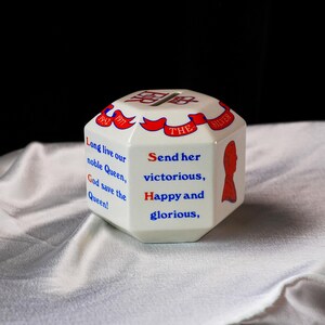 Op de afbeelding: Een witte, achthoekige keramische spaarpot met rode en blauwe tekst en accenten. De spaarpot heeft een gleuf voor munten en bevat de tekst "Long live our noble Queen, God save the Queen!" en "Send her victorious, Happy and glorious."