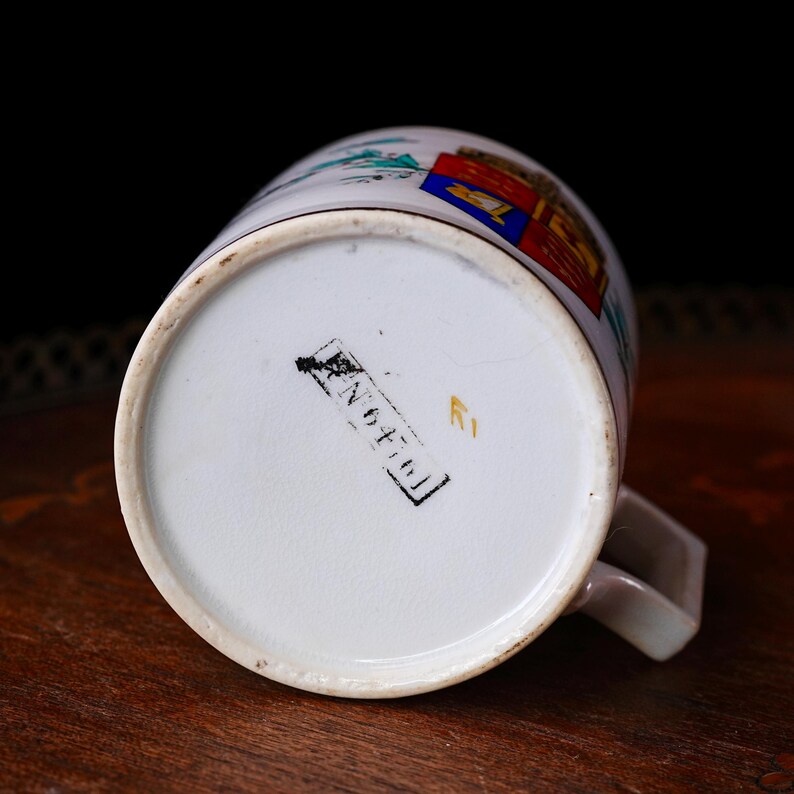 May include: White ceramic mug with a handle, featuring a colorful crest design and a black stamp with the text "N 6476". The mug's base is visible, showing a clean, white surface. The mug is set against a dark background.