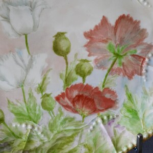 May include: A white ceramic plate with a hand-painted floral design featuring red and white poppies, green leaves, and seed pods. The plate has a scalloped edge with small, raised dots. The background is a soft pink and white.