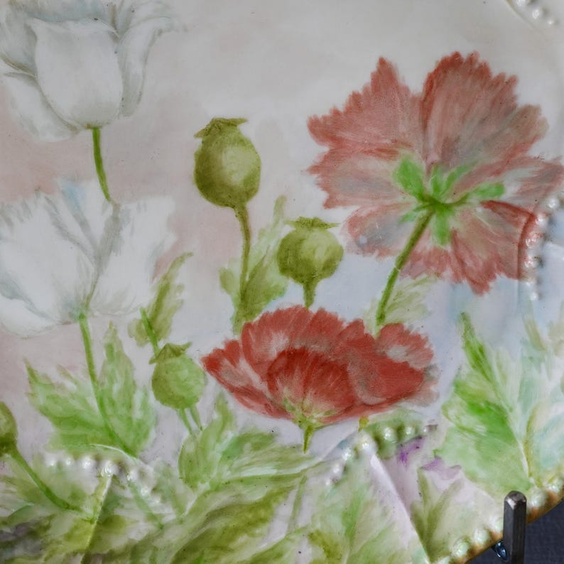 May include: A white ceramic plate with a hand-painted floral design featuring red and white poppies, green leaves, and seed pods. The plate has a scalloped edge with small, raised dots. The background is a soft pink and white.