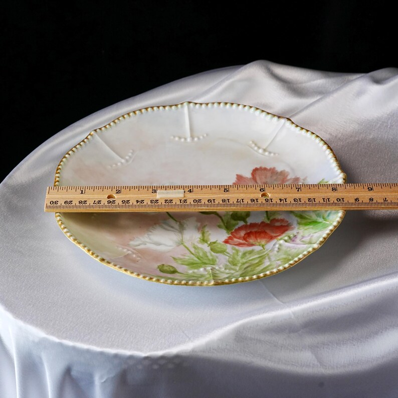 May include: Oval-shaped, antique porcelain plate with a hand-painted floral design featuring red poppies and green leaves. The plate has a scalloped edge with a gold beaded trim. The plate measures approximately 12 inches in length.