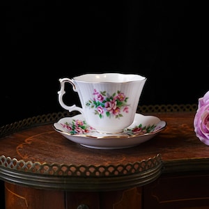 Op de afbeelding: Een witte theekop en schotel met roze en groene bloemmotieven en gouden randen. De set staat op een houten tafel met een decoratieve rand, met een roze roos op de achtergrond.
