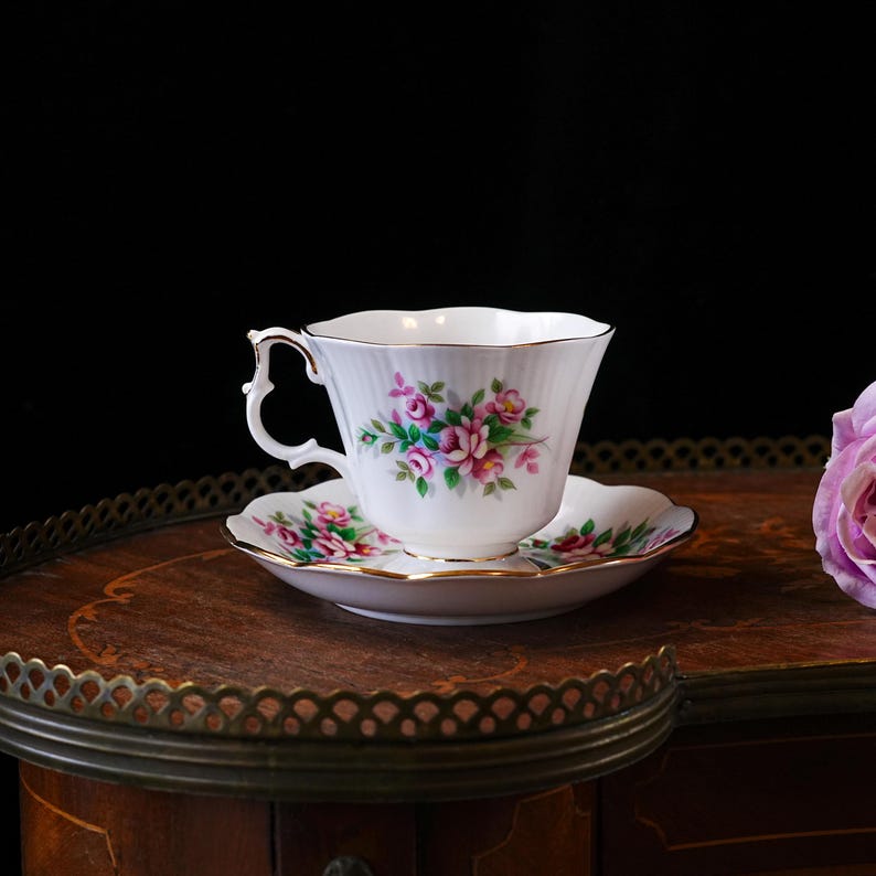Op de afbeelding: Een witte theekop en schotel met roze en groene bloemmotieven en gouden randen. De set staat op een houten tafel met een decoratieve rand, met een roze roos op de achtergrond.