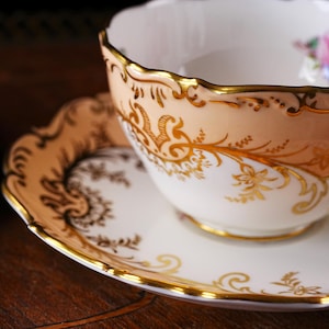 Op de afbeelding: Een vintage theekop en schotel set. De theekop heeft een perzikkleurige buitenkant met gouden filigraan en een geschulpte rand. De schotel is wit met een gouden rand en bijpassende gouden patronen. Een bloemenmotief is zichtbaar in de theekop.