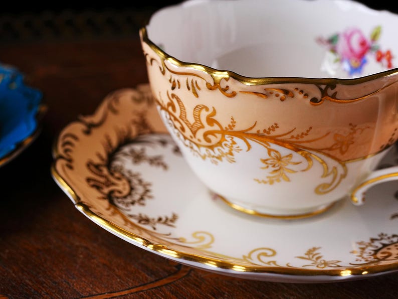Op de afbeelding: Een vintage theekop en schotel set. De theekop heeft een perzikkleurige buitenkant met gouden filigraan en een geschulpte rand. De schotel is wit met een gouden rand en bijpassende gouden patronen. Een bloemenmotief is zichtbaar in de theekop.