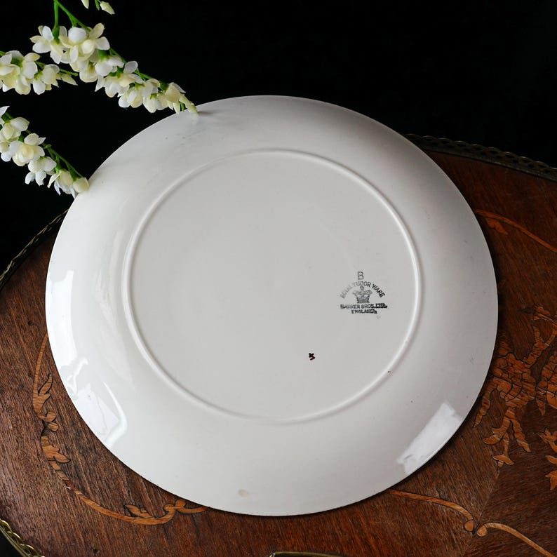 May include: A white ceramic plate with a wide rim, viewed from the underside. The plate has a stamp that reads "Royal Tudor Ware, Barker Bros Ltd, England." A small number "5" is visible. White flowers are in the upper left corner.