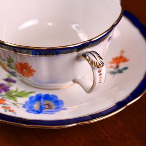 Puede incluir: Juego de taza y platillo blancos con dise&ntilde;o floral. La taza y el platillo tienen un borde azul con ribete dorado. El asa de la taza es blanca con detalles dorados. El dise&ntilde;o floral incluye flores naranjas, azules y moradas.