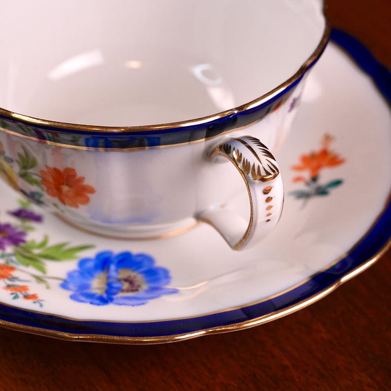 Puede incluir: Juego de taza y platillo blancos con dise&ntilde;o floral. La taza y el platillo tienen un borde azul con ribete dorado. El asa de la taza es blanca con detalles dorados. El dise&ntilde;o floral incluye flores naranjas, azules y moradas.
