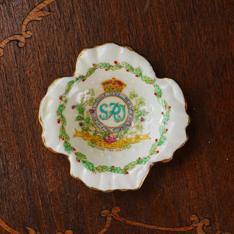 Op de afbeelding: Een kleine, witte, vierlobbige schaal met een gouden rand. De schaal heeft een centraal embleem met een kroon, de letters "SRJ" en de tekst "ET. SUON. DR. IT." omgeven door een bloemenkrans. De datum "MAY 12 1957" is ook zichtbaar.