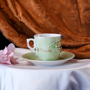 May include: A vintage teacup and saucer set. The cup and saucer are a pale green with gold accents and the words "A Present" in gold script. The cup has a decorative handle and floral embellishments.