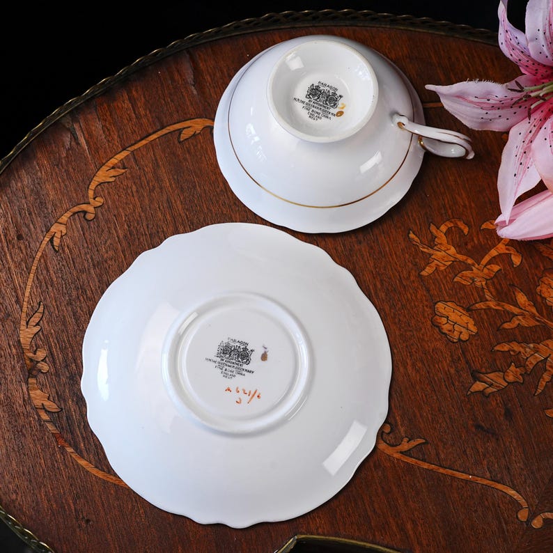 May include: A white porcelain teacup and saucer set featuring gold trim. The underside of both pieces displays a black maker's mark with the text PARAGON BY APPOINTMENT FINE BONE CHINA ENGLAND. The saucer also features handwritten red text.