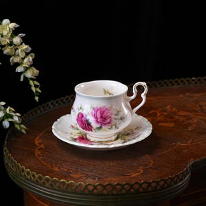 May include: A fine bone china teacup and saucer, decorated with pink rose motifs. The cup has a delicate heart-shaped handle. The set is displayed on a dark, ornate wooden table, showcasing a classic design.