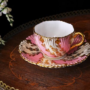 May include: A pink and gold teacup and saucer set. The teacup and saucer have a delicate floral design with gold trim. The set is placed on a dark wooden tray with a decorative edge. White flowers are in the background.
