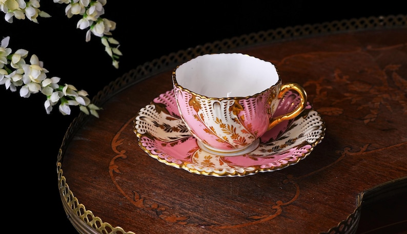 May include: A pink and gold teacup and saucer set. The teacup and saucer have a delicate floral design with gold trim. The set is placed on a dark wooden tray with a decorative edge. White flowers are in the background.