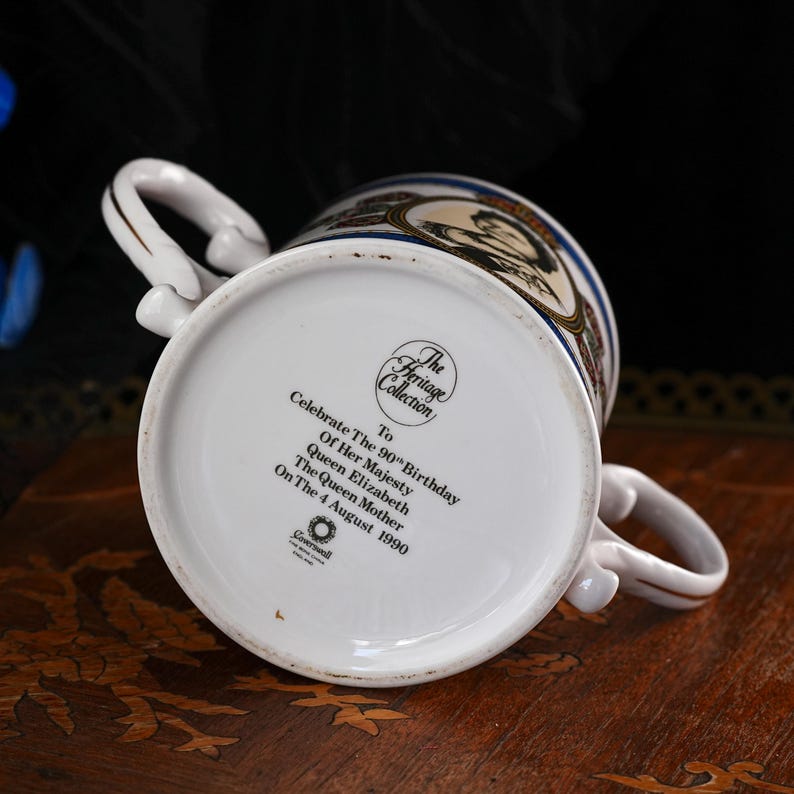 May include: A white porcelain two-handled loving cup featuring a commemorative print on the base that reads: The Heritage Collection, To Celebrate The 90th Birthday Of Her Majesty Queen Elizabeth The Queen Mother On The 4 August 1990, Coalport.