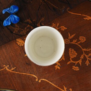 Puede incluir: Una taza de cer&aacute;mica blanca con un borde dorado vista directamente desde arriba. El interior de la taza es liso y de color blanco roto con peque&ntilde;as manchas oscuras visibles en la base. La taza tiene forma cil&iacute;ndrica y un borde dorado fino.