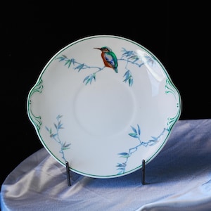 May include: White ceramic plate with a green rim and handles. The plate features a colorful illustration of a bird perched on a branch with blue leaves. The plate is displayed on a black metal stand.