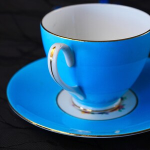 Op de afbeelding: Een fel turquoise theekop en schotel set met een witte binnenkant en een fijne gouden rand langs de rand en het handvat. Het handvat is wit met een gouden streep en het midden van de schotel toont een klein, kleurrijk bloemmotief.