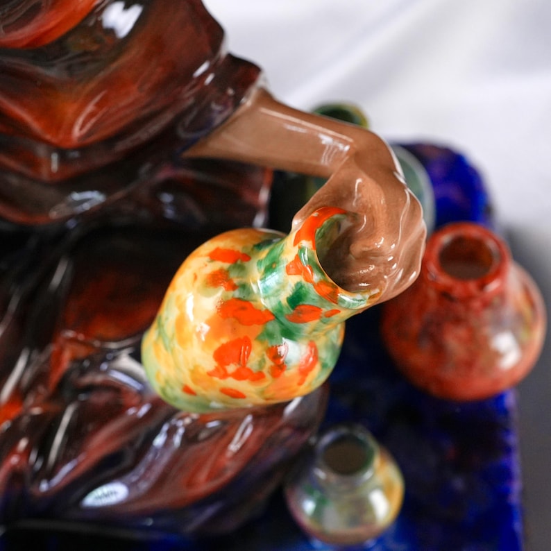 May include: A ceramic figurine of a person holding a small, colourful vase. The vase is yellow with orange and green spots. Other small vases in red, green, and blue are also visible. The figurine has a dark red and brown colour scheme.